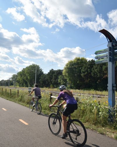 Two people ride bicycles on a paved trail next to train tracks, surrounded by greenery and a signpost under a partly cloudy sky.